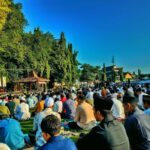 Large crowd seated on prayer mats for an outdoor communal prayer in sleman with trees and a stage in the background